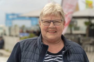 Lächelnde ältere Frau mit Brille und kurzen grauen Haaren, trägt eine dunkelblaue Jacke und ein schwarz-weiß gestreiftes Oberteil, draußen an einem Hafen mit Booten im Hintergrund.
