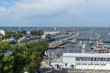 Blick auf einen Hafen mit mehreren Segelbooten auf dem Wasser, ein Leuchtturm im Hintergrund, und umgeben von Gebäuden und Bäumen, bei überwiegend bewölktem Himmel.