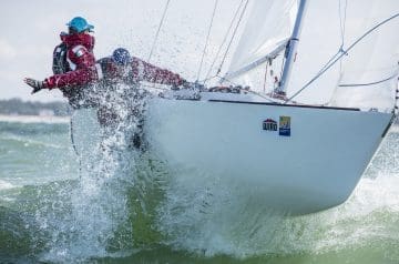 Segler in roter Kleidung mit italienischer Flagge auf dem Arm, der auf einem weißen Segelboot bei windigem Wetter auf dem Wasser segelt, während das Boot Wasser spritzt.