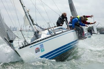 Menschen auf einem segelboot, das gekippt ist, bei einem Rennen im Wasser, mit mehreren Personen, die das Gleichgewicht halten, während sie das Boot steuern, unter bewölktem Himmel.