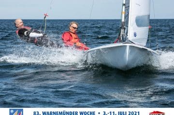 Zwei Menschen in einem weißen Segelboot auf dem Meer während eines Segelrennens, beide tragen rote Weste, eine Person liegt im Wasser neben dem Boot, während die andere im Boot sitzt, die Wellen sind sichtbar, Himmel ist klar.