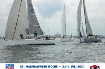 Mehrere Segelboote auf dem Wasser während eines Regattalaufs vor einer Küstenstadt, mit einem Wolkenverhangenen Himmel im Hintergrund, bei der Warnemünder Woche im Juli 2021.