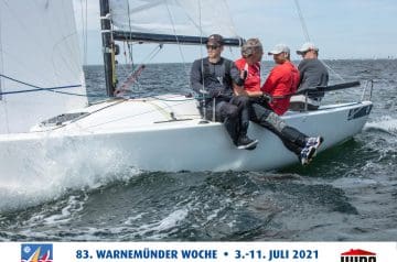 Vier Männer sitzen auf einem weißen Segelboot auf dem Wasser, eines trägt schwarze Kleidung und trägt Sonnenbrille, während die anderen in roten und grauen Kleidern sind und eine Diskussion führen. Das Boot ist bei einer Segelregatta, mit mehreren anderen Segelbooten im Hintergrund.