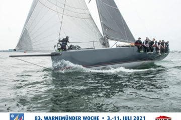 Ein Segelboot mit mehreren Personen an Bord bei einer Regatta, das auf dem Wasser treibt, mit großen Segeln und einer grauen Schiffshaut, bei bewölktem Himmel.