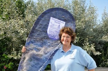 Eine lachende Frau mit brünetischem, lockigem Haar trägt ein hellblaues Hemd mit dem Logo 'Warnemünde Verein' und steht vor grünen Büschen. Sie hält ein großes Flaggenbanner mit Wasserwellen-Design und dem Logo des Vereins hoch.