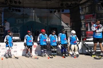 Hockeyspieler in blau-weißen Trikots mit Helm und Schlägern stehen in einer Reihe auf einem Spielfeld, ausgestattet mit Schutzkleidung, auf dem Boden liegen Hockeybälle. Ein Trainer oder Betreuer steht rechts, trägt Skates, ein blauen Trikot und rote Handschuhe.