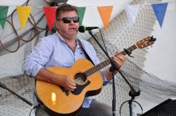 Ein Mann mit Kurzhaar, Bart und Sonnenbrille spielt auf einem Akustik-Gitarre, während er vor einem Mikrofon singt bei einem festlichen Anlass mit bunten Wimpeln im Hintergrund.
