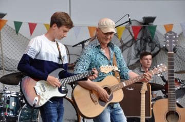 Zwei junge Männer und ein älterer Mann mit langem Haar spielen Gitarre und E-Gitarre auf einer Bühne, hinter ihnen bunte Fähnchen und Musikinstrumente, während sie sich beim Musizieren konzentrieren.