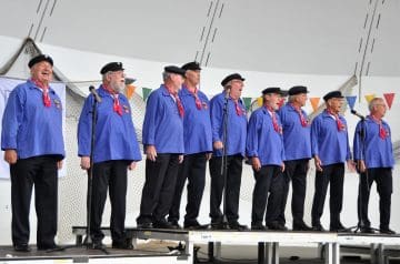 Eine Männergruppe steht auf einer Bühne und singt, alle tragen blaue Hemden, schwarze Hüte und rote Halstücher, im Hintergrund sind bunte Fahnen und ein Netzartige Dekoration sichtbar.