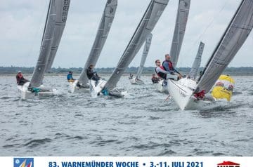 Mehrere Segelboote bei einer Regatta auf dem Wasser, mit einer Gruppe von Seglern am Steuer, und eine gelbe Boje im Wasser, bei bewölktem Himmel, im Hintergrund eine flache Küstenlinie.