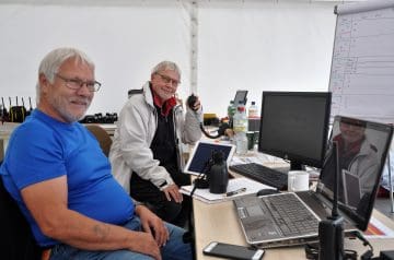 Zwei ältere Männer mit Brillen sitzen an einem Tisch in einem Raum, einer trägt ein blaues T-Shirt, der andere eine weiße Jacke, und beide schauen in die Kamera, während auf dem Tisch technische Geräte und Wasserflaschen stehen.