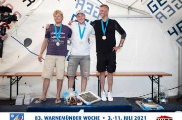 Drei Männer auf einem Siegerpodest bei einer Regatta, die Medaillen um den Hals tragend, lächelnd und umarmend. Im Hintergrund eine weiße Zeltwand mit blauen und schwarzen Schriftzügen.