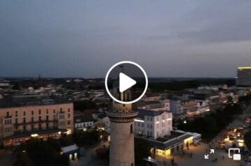 Luftaufnahme einer Stadt bei Abenddämmerung, im Vordergrund ein hoher, runder Turm mit fransenförmigem Balkon, im Hintergrund moderne Gebäude und eine dunkle Wolkendecke.