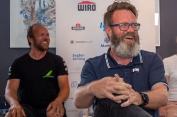 Zwei Männer lachen und sitzen in einem Raum mit Logos im Hintergrund, einer trägt eine schwarze T-Shirt und der andere eine dunkelblaue Poloshirt mit Brille und Bart.