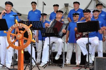 Eine Gruppe von Männern in blauen Poloshirts und weißen Hosen spielt auf einer Bühne, wobei einer Akkordeon spielt, während andere Gitarre spielen und singen, mit einem Steuerrad mit der Aufschrift "DE KLAASHAVENS" vor ihnen.