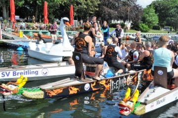 Boot mit Drachendekorationen trägt Menschen, die an einer Feier oder Veranstaltung auf dem Wasser teilnehmen, mit einem Publikum im Hintergrund.