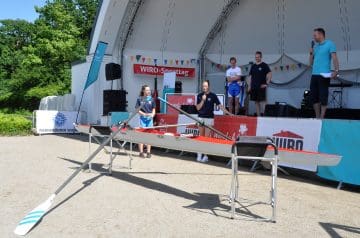 Ein Outdoor-Bühnenszene bei einem Wassersport-Event mit mehreren Personen, einem Surfbrett und einem Paddel im Vordergrund, während im Hintergrund eine Bühne mit Banner, Redner und Zuschauern steht.