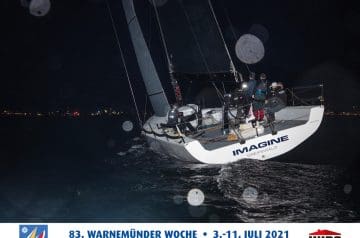 Segelboot bei Nacht auf dem Wasser mit mehreren Crewmitgliedern an Bord, das Wasser ist dunkel und der Himmel wolkenbedeckt, im Hintergrund sind Stadtlichter sichtbar