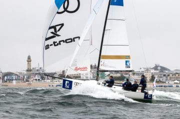 Ein Segelboot mit vier Personen, die bei windigem Wetter auf dem Wasser segeln, vor einer Stadtansicht mit Booten, Gebäuden und einem Leuchtturm im Hintergrund