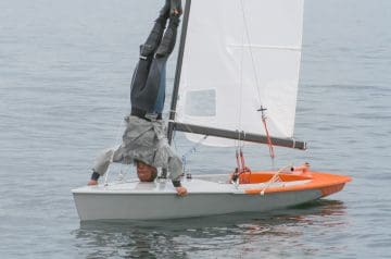 Ein Mann liegt kopfüber auf einem kleinen Segelboot auf dem Wasser, hält sich mit den Händen am Boot fest und hat die Beine in die Luft gestreckt, während die Segel entfaltet sind.