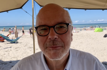 Ein älterer Mann mit Brille und weißem Bart sitzt unter einem Sonnenschirm am Strand, im Hintergrund sind Menschen, die im Sand liegen, im Wasser schwimmen und am Strand spazieren gehen, bei gutem Wetter mit blauer Himmel und wenigen Wolken.