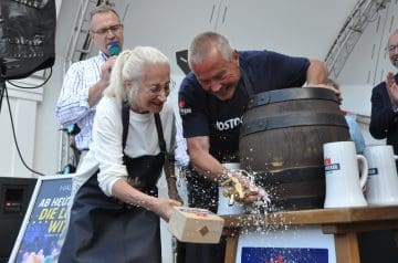 Mehrere Menschen, darunter ein älterer Frau und ein Mann, die ein Holzfass mit einem Hammer zerschlagen, während Wasser oder Saft spritzt, bei einer Veranstaltung. Es ist während eines Auftritts oder Festes, mit anderen Personen im Hintergrund.