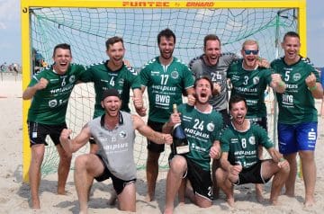 Eine Gruppe fußballspieler in grünen Trikots jubelt vor einem Beachsoccer-Tor am Strand, einige knien und einige stehen, während sie eine Flasche Sekt halten und vor Freude lachen.