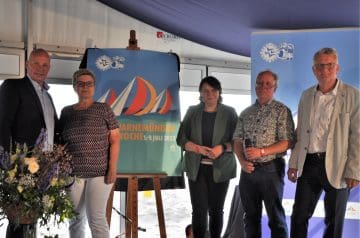 Fünf Menschen stehen nebeneinander vor einer blauen Bannerwand bei einer Veranstaltung, im Hintergrund sind Sonnenlicht und das Meer sichtbar.