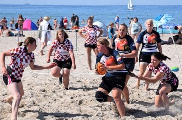 Eine Gruppe von Frauen spielt Beach-Rugby am Strand, mit einigen im Hintergrund, während das Meer und Boote zu sehen sind.