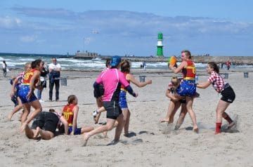 Menschen spielen Beach Rugby am Strand, einige Frauen in roten, weißen und blauen Trikots, eine Frau springt mit dem Ball, im Hintergrund ein Leuchtturm und die See.