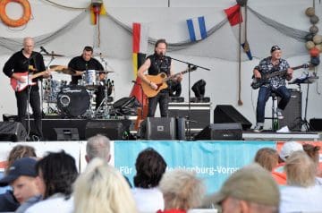 Eine Band mit vier männlichen Musikern spielt auf einer Bühne vor einem Publikum. Der Musiker in der Mitte singt und spielt Gitarre, die anderen spielen E-Gitarre, Schlagzeug und Bass. Die Bühne ist mit Flaggen dekoriert, darunter Deutschland, Griechenland und Finnland, und es gibt Lautsprecher und technische Ausrüstung.