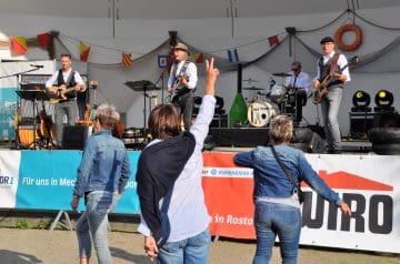 Eine lebendige Outdoor-Bildaufnahme einer Band, die auf einer Bühne auftritt, während mehrere Frauen im Vordergrund tanzend und zuschauend sind, mit bunten Flaggen und einem Werbebanner im Hintergrund.