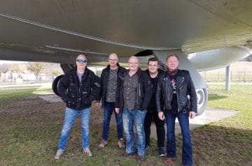 Fünf Männer stehen vor einem großen Flugzeug, das auf einer Wiese geparkt ist, und posieren für ein Foto. Sie tragen alle Jacken und jeans und scheinen sich zu amüsieren.