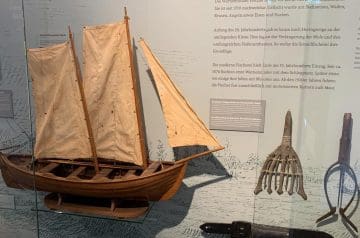 Modell eines historischen Ruderboots mit hellen Segeln neben verschiedenen alten Fischerwerkzeugen auf einer Ausstellung.