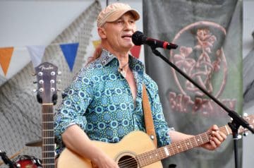 Ein älterer Mann spielt Gitarre und singt in einem Mikrofon auf einer Bühne, im Hintergrund sind bunte Fahnen und ein Banner mit einem roten Logo. Er trägt ein beiges Cap und ein blau-mustertes Hemd und hat längliches, rotes Haar.