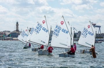 Mehrere Optimist-Segelboote mit deutschen Kennzeichnungen segeln auf dem Wasser, während sie an einem Wettkampf teilnehmen, mit einer Küstenstadt im Hintergrund und einem bewölkten Himmel.