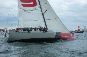 Ein Segelboot mit mehreren Crewmitgliedern, das auf dem Wasser fährt, mit großen weißen Segeln und der Aufschrift "Echt von hier. Nah bei mir. Meine OSPA." am Heck, während im Hintergrund ein Leuchtturm und ein Riesenrad zu sehen sind.