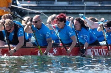 Mehrere Menschen beim Drachenbootrennen, die in einem roten Boot paddeln, tragen blaue T-Shirts und paddeln im Wasser.