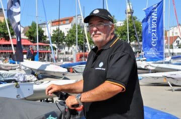Ein älterer Mann mit Brille, grauem Bart und grauen Haaren, trägt eine schwarze Polo-Hemd mit orangefarbenem Rand an den Ärmeln und einem schwarzen Cap, der auf einem Bootssteg bei Segelbooten steht.