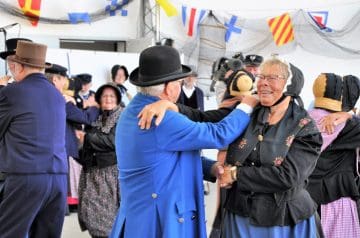 Ältere Menschen in traditioneller Kleidung tanzen auf einer festlichen Veranstaltung, im Hintergrund bunte Flaggen und Netzdekorationen. Ein Mann in blauem Anzug und Hut hält eine Frau mit Brille, Kopfbedeckung und schwarzer Kleidung, beide lachen.