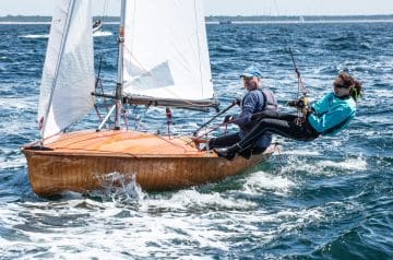 Zwei Menschen auf einem kleinen Segelboot, das auf dem Wasser fährt, mit weißen Segeln und einer Holzkonstruktion, unter sonnigem Himmel.