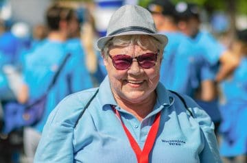 Eine ältere Frau trägt einen grauen Hut, große Sonnenbrille und ein hellblaues Hemd mit dem Schriftzug 'Wornemünde Verein', sie lächelt. Im Hintergrund sind mehrere Menschen in blauen T-Shirts bei einer outdoor Veranstaltung, der Hintergrund ist unscharf.