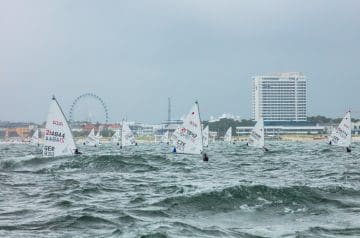 Mehrere Windsurfer auf dem Wasser vor einer Küstenstadt mit hohen Gebäuden, einem Riesenrad und einer belebten Strandpromenade bei windigem Wetter.