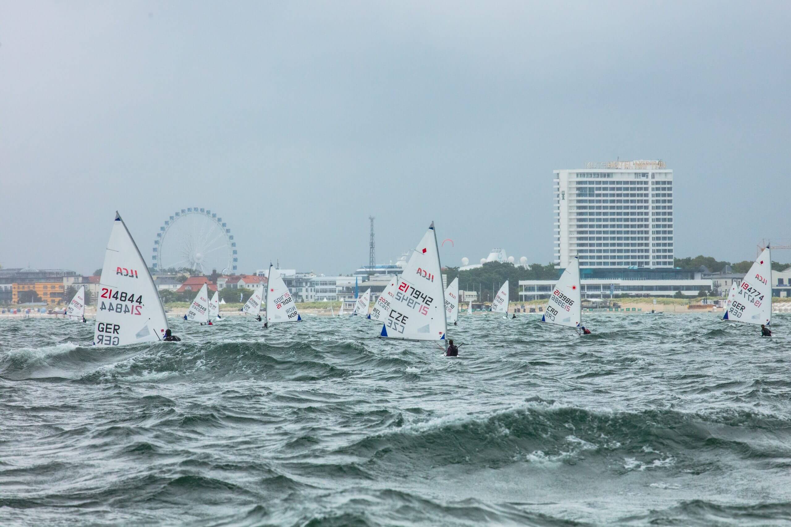 Mehrere Windsurfer auf dem Wasser vor einer Küstenstadt mit hohen Gebäuden, einem Riesenrad und einer belebten Strandpromenade bei windigem Wetter.