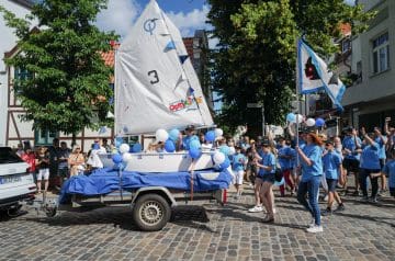 Eine bunte Parade mit Menschen in blauen T-Shirts, die einen kleinen Segelboot-Umzug auf einer cobbelstonierten Straße veranstalten, begleitet von Zuschauern und Fahnen bei schönem Wetter.