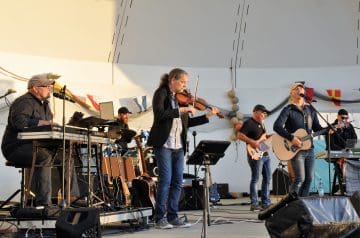 Eine Live-Band spielt auf einer Bühne, bestehend aus mehreren Musikern, darunter ein Violinist, Sänger mit Gitarre, E-Gitarrist, Bassist und Keyboarder, vor einer weißen Wand mit bunten Stoffdrapierungen und Dekorationen.