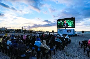 Menschen sitzen in Stühlen und schauen einen großen Bildschirm, der einen Vortrag oder Show zeigt, am Hafen bei Sonnenuntergang mit Booten und Gebäuden im Hintergrund.