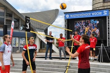 Volleyballspieler in roten und weißen Trikots spielen im Freien bei einem Event, während ein Zuschauer den Ball hochwirft und eine Großbildleinwand im Hintergrund eine Gruppe von Menschen zeigt, die die Spielszenen verfolgen. Es gibt eine Bühne mit einigen Moderatoren, ein Banner mit der Aufschrift "WARNEMÜNDER WOCHE" und eine moderne Architektur im Hintergrund.