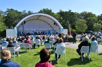 Eine Gruppe von Menschen sitzt im Freien vor einer kleinen Bühne im Park, während eine Veranstaltung mit Musik oder Reden stattfindet, umgeben von Bäumen bei sonnigem Wetter.