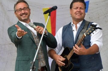 Zwei Männer auf einer Bühne, einer hält ein Mikrofon und trägt eine Brille, der andere spielt Gitarre, beide tragen formelle Kleidung und scheinen bei einer Veranstaltung aufzutreten.
